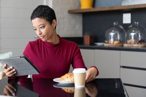 African american woman reading tablet while enjoying coffee and croissant in kitchen cafe