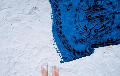 Top-down showing bare feet on white sand beside blue tropical sarong