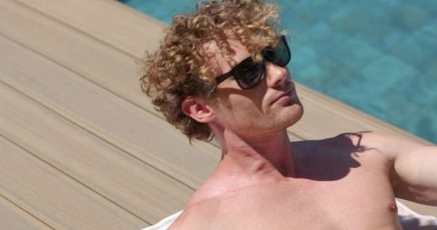 Young man relaxing shirtless on pool deck wearing sunglasses by turquoise water, sunny