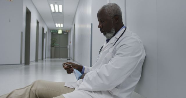 Black male doctor sitting alone on hospital floor, holding mask and reflecting on workload. Useful for depicting healthcare challenges, doctor fatigue, medical professional stress, emotional impact of healthcare jobs, crisis situations, and importance of mental health support in medical field.