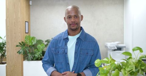 Standing professional wearing denim shirt in modern office with plants and multifunction printer