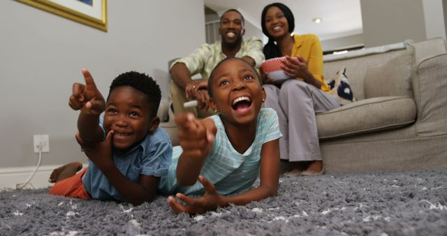 African American family enjoying time together, with the children lying on the carpet and pointing at the camera, and the parents sitting on the couch smiling, with copy space. Their joyful expressions and the cozy home setting convey a sense of warmth and family bonding.