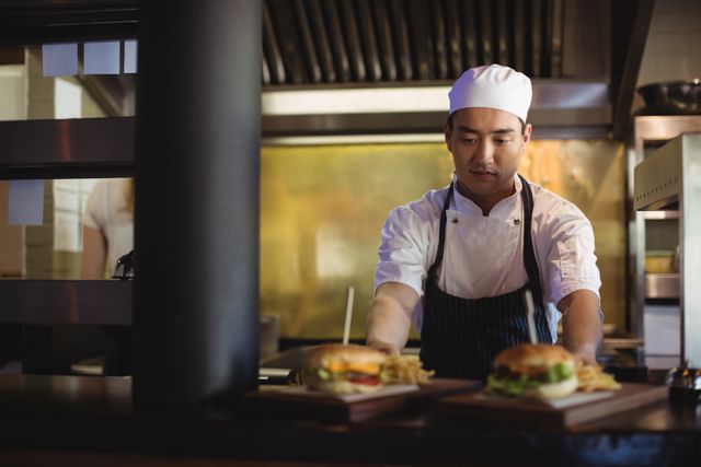 Chef wearing white uniform and hat, placing tray with burgers and french fries at order station in busy commercial kitchen. Ideal for use in content about restaurant industry, professional cooking, culinary education, or fast food services.