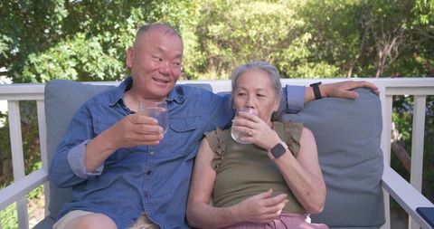 Asian senior couple relaxing on porch bench, sipping water and enjoying sunlit garden