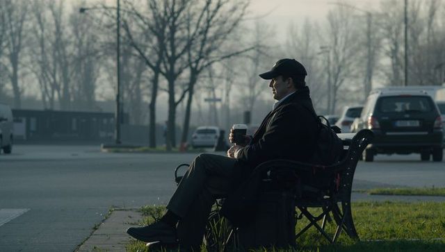 Solitary traveler sitting on bench holding to-go coffee by roadside in misty urban morning