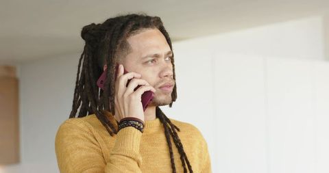 Young man with dreadlocks making phone call at home wearing mustard sweater and bracelets