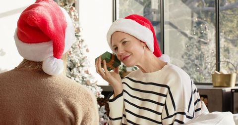 Two friends wearing santa hats exchanging gift by christmas tree in cozy holiday home