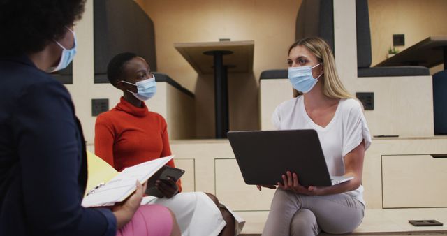 Three two diverse female office colleagues wearing face masks discussing together at office. hygiene and social distancing in the workplace during coronavirus covid 19 pandemic.