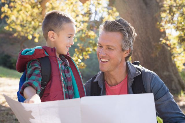 Father and son sharing a joyful moment while looking at a map in a forest setting. Ideal for themes related to family bonding, outdoor activities, and adventure. Can be used in marketing materials for travel agencies, outdoor gear, or family-focused content.