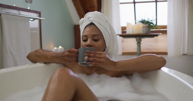Image of a woman enjoying a relaxing bubble bath while drinking tea in a cozy bathroom. Useful for promoting luxury bath products, wellness and self-care routines, home improvement, and stress-relief services.