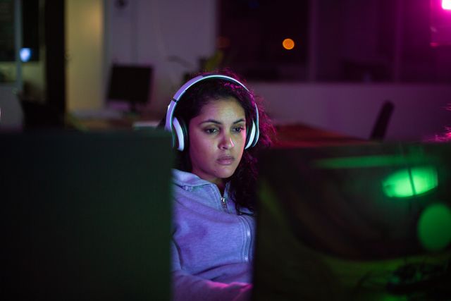Woman working late in a creative office, wearing headphones and typing on a computer keyboard. Ideal for illustrating concepts of dedication, late-night work, technology use, and modern office environments.