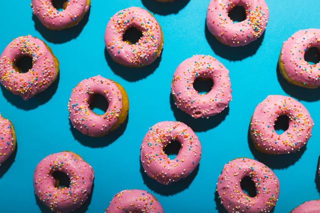 Directly above view of fresh pink donuts with sprinklers arranged in row against blue background. unaltered, unhealthy eating and sweet food concept.