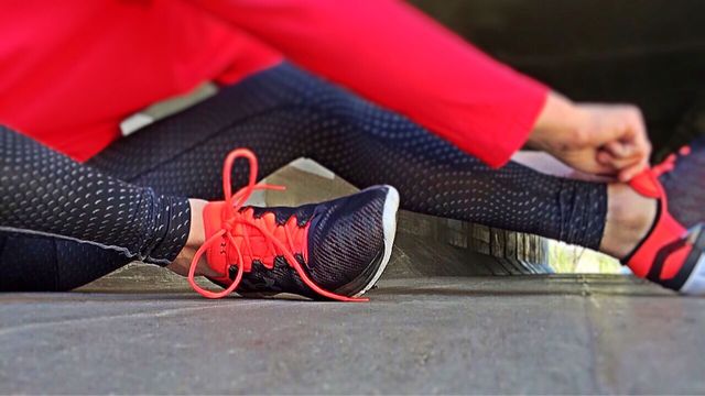 Female athlete tying bright red running shoes while preparing for exercise. She is wearing patterned leggings and red sportswear on concrete surface. Ideal for fitness, workout, or athletic-themed content.