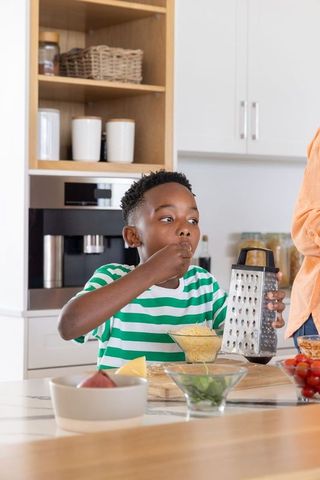 African american child tasting grated cheese holding metal box grater at kitchen island