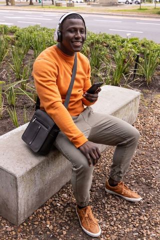 African american man smiling, listening to music on headphones while holding smartphone