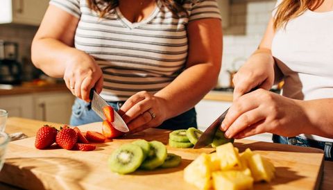Two women chopping strawberries kiwi pineapple on wooden board in bright home kitchen