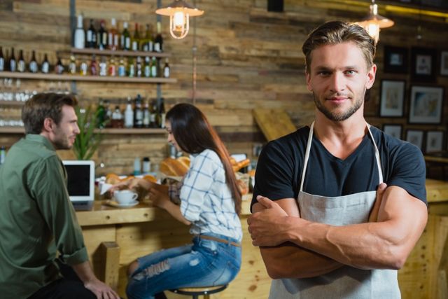 Confident waiter standing with arms crossed in a trendy café with wooden interior. Two customers are sitting at the counter, engaging in conversation. Ideal for use in articles about the hospitality industry, small businesses, café culture, or customer service.