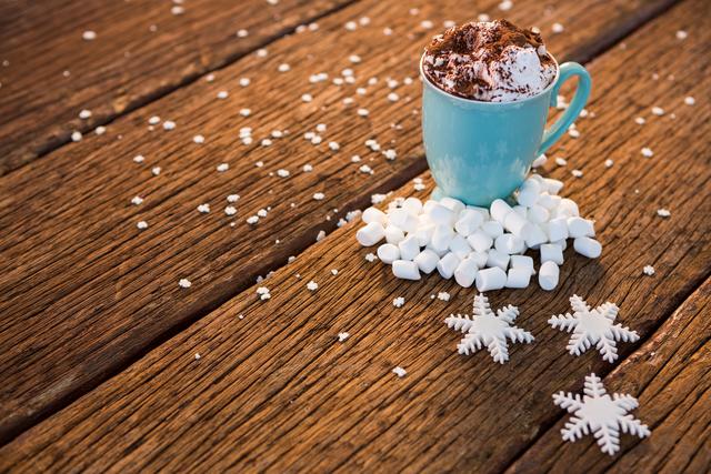Warm cup of hot chocolate topped with cocoa powder and surrounded by marshmallows and snowflake ornaments on a rustic wooden surface. This image evokes a cozy and festive winter atmosphere, perfect for holiday-themed content, Christmas greetings, or winter drinks promotions.