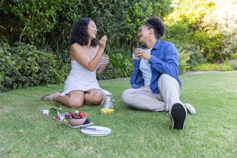 Diverse couple enjoying sunny garden picnic, laughing and sharing fresh fruit
