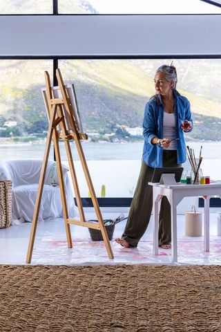Senior asian woman painting at easel in bright waterfront studio, holding brush and glass