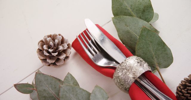 A festive table setting features a silver fork and knife wrapped in a red napkin, adorned with green leaves and pine cones, with copy space. It evokes a cozy, holiday atmosphere, ideal for seasonal celebrations.