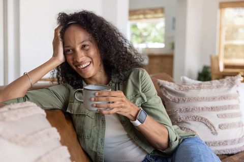 Relaxed african american woman holding mug wearing smartwatch on couch in modern living room