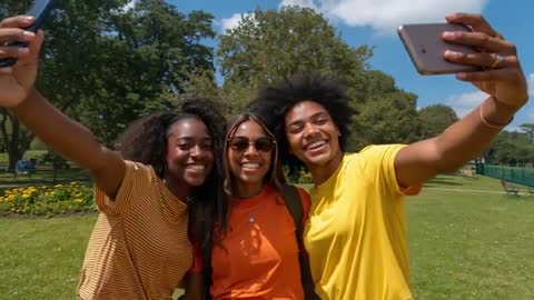 Young friends taking dual-phone selfie in sunny park, laughing and posing for camera
