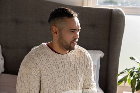 Mid-adult asian man sitting on bed wearing cream cable-knit sweater by sunlit window