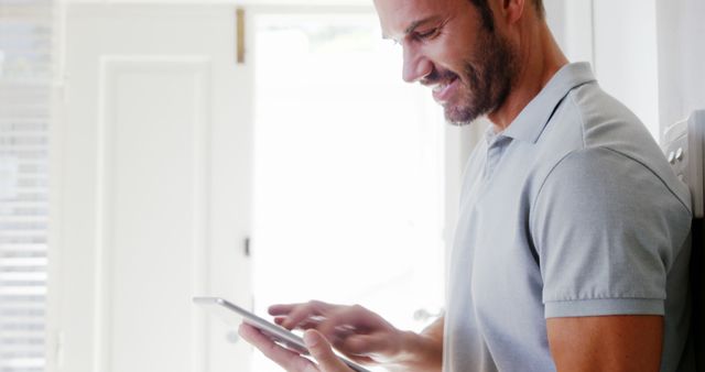 Man standing indoor wearing casual shirt, using digital tablet, cheerful expression. Ideal for illustrating concepts related to technology use in daily life, online activities, or remote working scenarios.