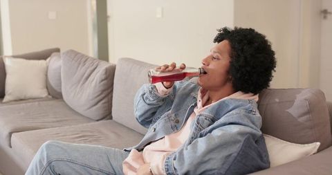 African american man relaxing on sofa drinking bottled beverage in denim jacket and hoodie