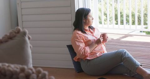Mature woman relaxing on floor by sliding door holding mug, morning porch reflection calm