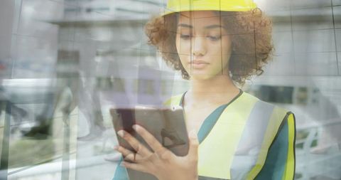 Female inspector reviewing tablet through glass in hard hat and hi-vis vest urban reflection