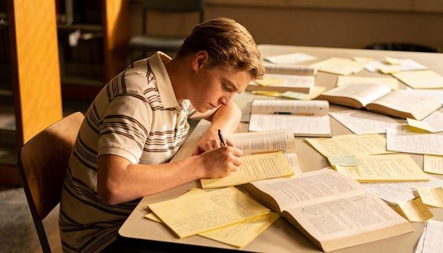 Focused teen studying at library table writing notes on yellow pad for exam prep