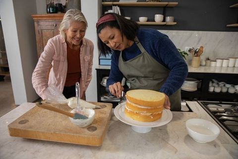 Mature women assembling three-layer cake in kitchen, frosting together with piping bag