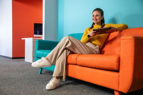 Young woman relaxing on orange sofa holding tablet wearing smartwatch in modern office
