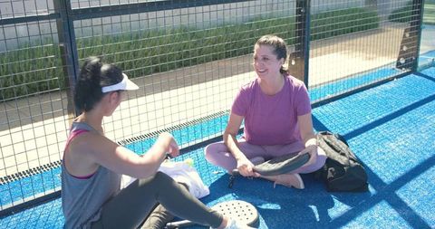 Women sitting and laughing after padel match on bright blue outdoor court with paddles