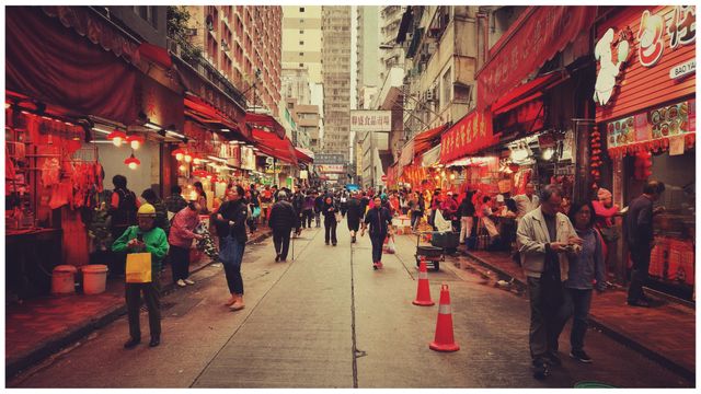 Busy Asian street market scene with several vendors selling goods to local shoppers. The market has vibrant red hues, traditional lanterns, and bustling activity. Ideal for illustrating cultural immersion, urban commerce, or as a background for travel and city living themes.