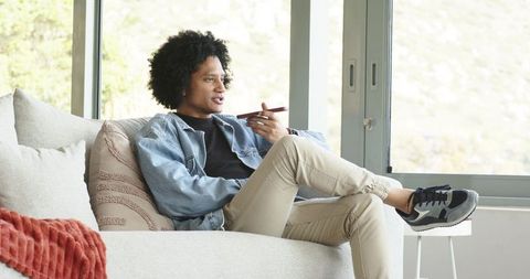 African american man relaxing on modern sofa speaking into smartphone in bright lounge