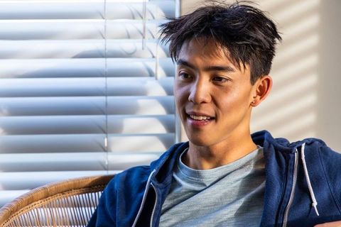 Sunlit asian man relaxing in rattan chair by blinds, casual navy hoodie lifestyle portrait