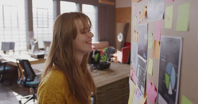 Young Caucasian woman reviews a colorful storyboard in an office. Her focus and smile suggest a creative brainstorming session at work.