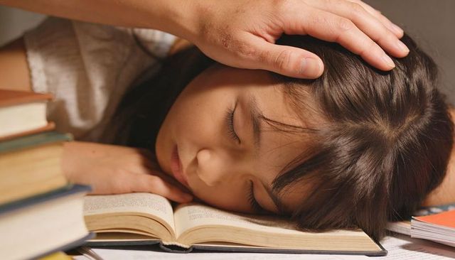 Young girl sleeping on open book with caring hand over head cozy study nap at home desk