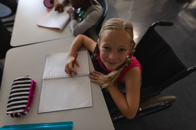 Young schoolgirl in a wheelchair smiling while writing in a notebook at her desk in a classroom. Ideal for use in educational materials, inclusive education campaigns, and promoting diversity and accessibility in schools.