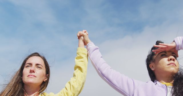 Two women wearing casual jackets holding hands outdoors beneath a bright blue sky. One woman is looking forward confidently, and the other is shielding her eyes from the sun with her free hand. This image can be used to represent concepts of friendship, unity, empowerment, support, teamwork, and a positive connection between people.