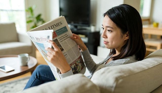Woman reading newspaper on sofa at home morning routine with coffee nearby