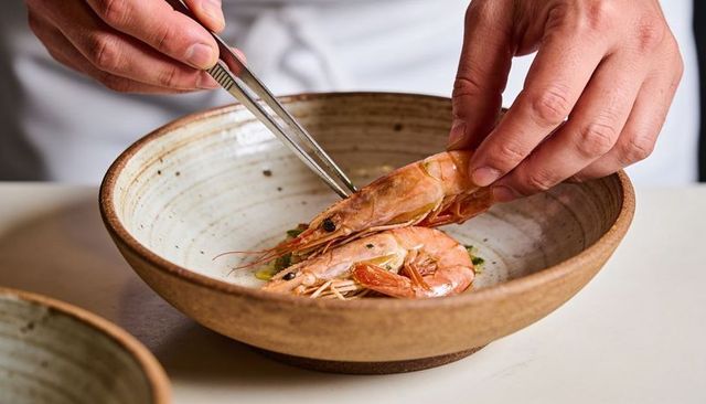 Chef plating whole prawns with tweezers in rustic ceramic bowl for elegant seafood presentation