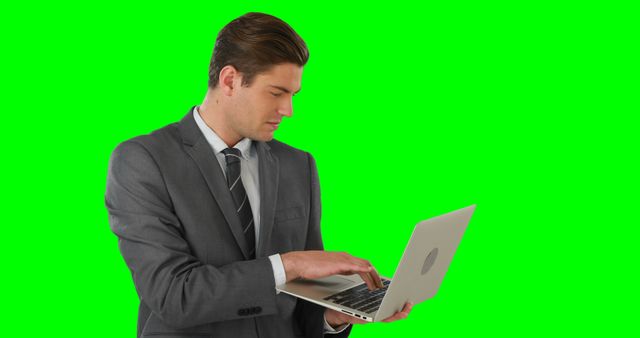 Businessman in a gray suit typing on a laptop against a chroma key green background. This can be used in business and technology presentations, promotional materials for corporate environments, or any project requiring the depiction of remote work or professional settings.