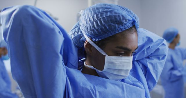 Biracial female surgeon putting on protective clothes standing in operating theatre. medicine, health and healthcare services.
