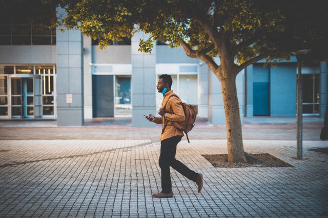 African American man wearing face mask and backpack walking in urban area while using smartphone. Ideal for illustrating themes of urban lifestyle, business on the go, pandemic safety measures, and modern city living.