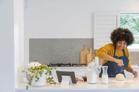 African american woman kneading dough in bright kitchen with tablet and flour jar