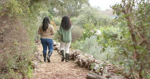 Diverse women walking woodchip trail in sweaters and rubber boots through lush garden